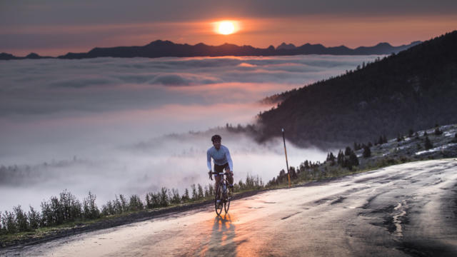 Une cycliste au sommet d'un col avec vue sur le coucher de soleil et les montagnes